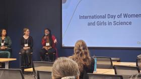 Photo of three women sitting at the front of a meeting room with a large screen that says "International Day of Women and Girls in Science." One woman who is African American, holds a mic while she is speaking.