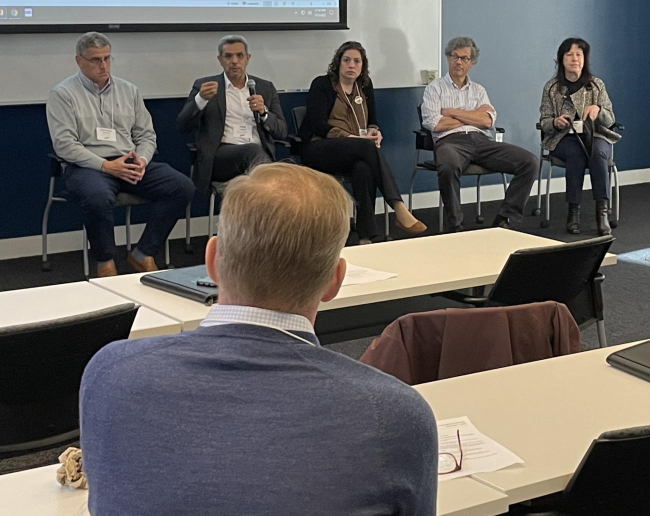 Photo of five McCamish conference panelists. Four panelists (one man and three women) listen to the fourth participant, a male, speaking.